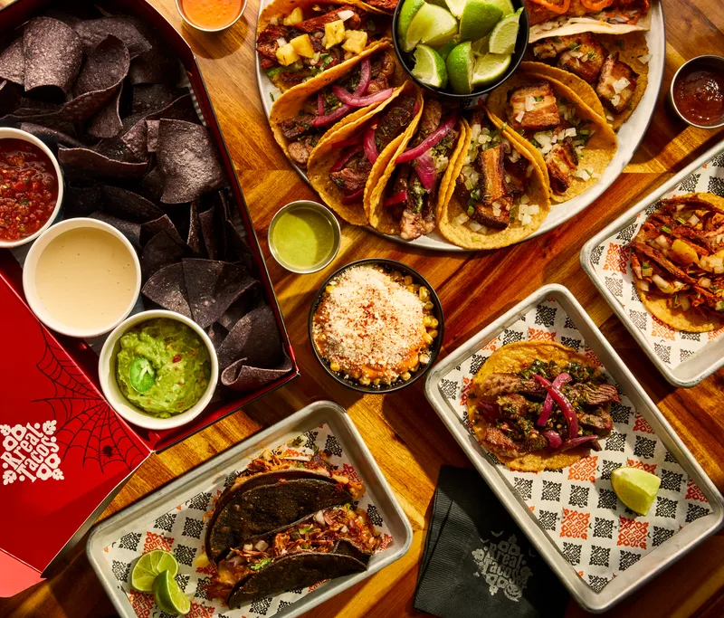 Open Rreal Tacos Halloween takeout box — “Haunted by Rreal Tacos” — with chips, salsa, queso and guacamole, next to tacos