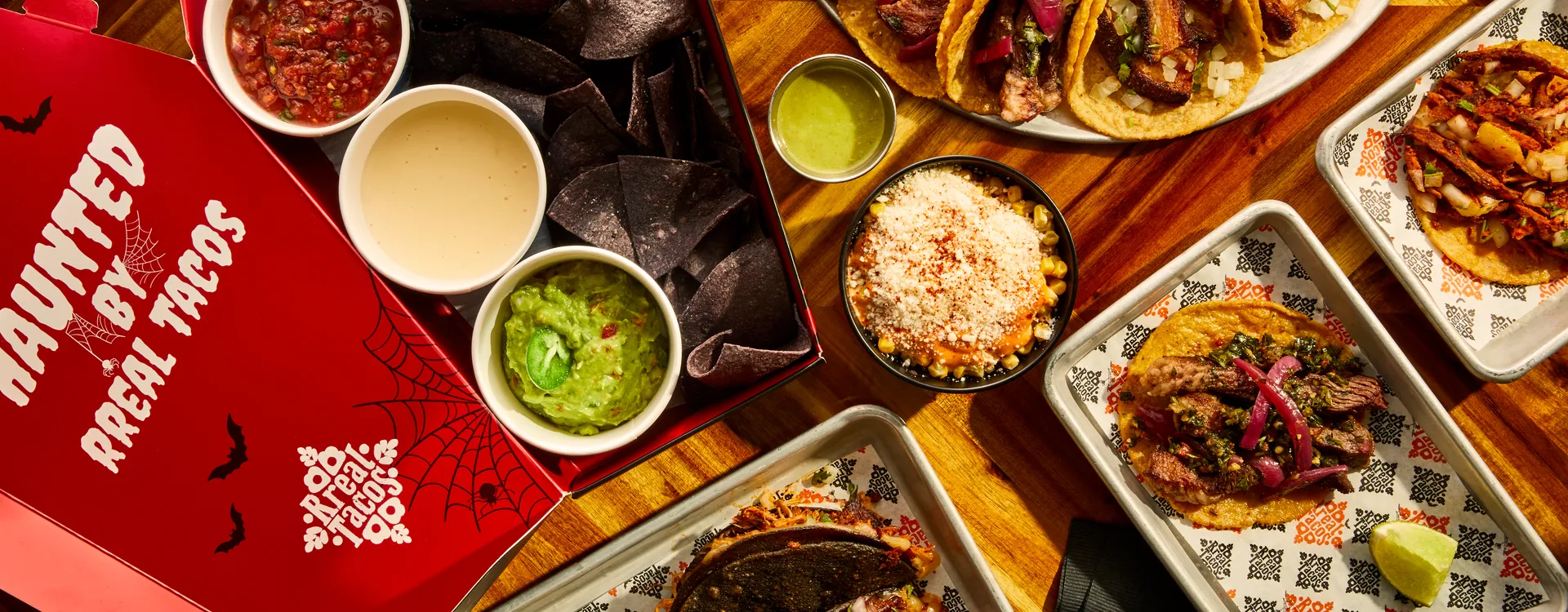 Open Rreal Tacos Halloween takeout box — “Haunted by Rreal Tacos” — with chips, salsa, queso and guacamole, next to tacos