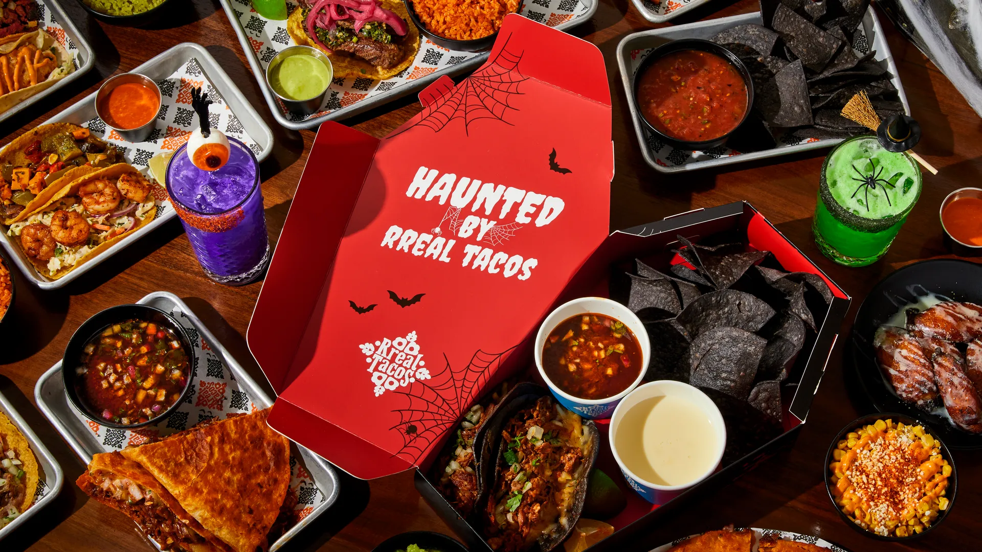 Rreal Tacos Halloween takeout box — “Haunted by Rreal Tacos” — surrounded by tacos, chips and themed drinks